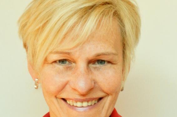 Porträt einer lächelnden Frau mit blonden Haaren, die ein schwarz-rotes Oberteil trägt, vor einem hellen Hintergrund.